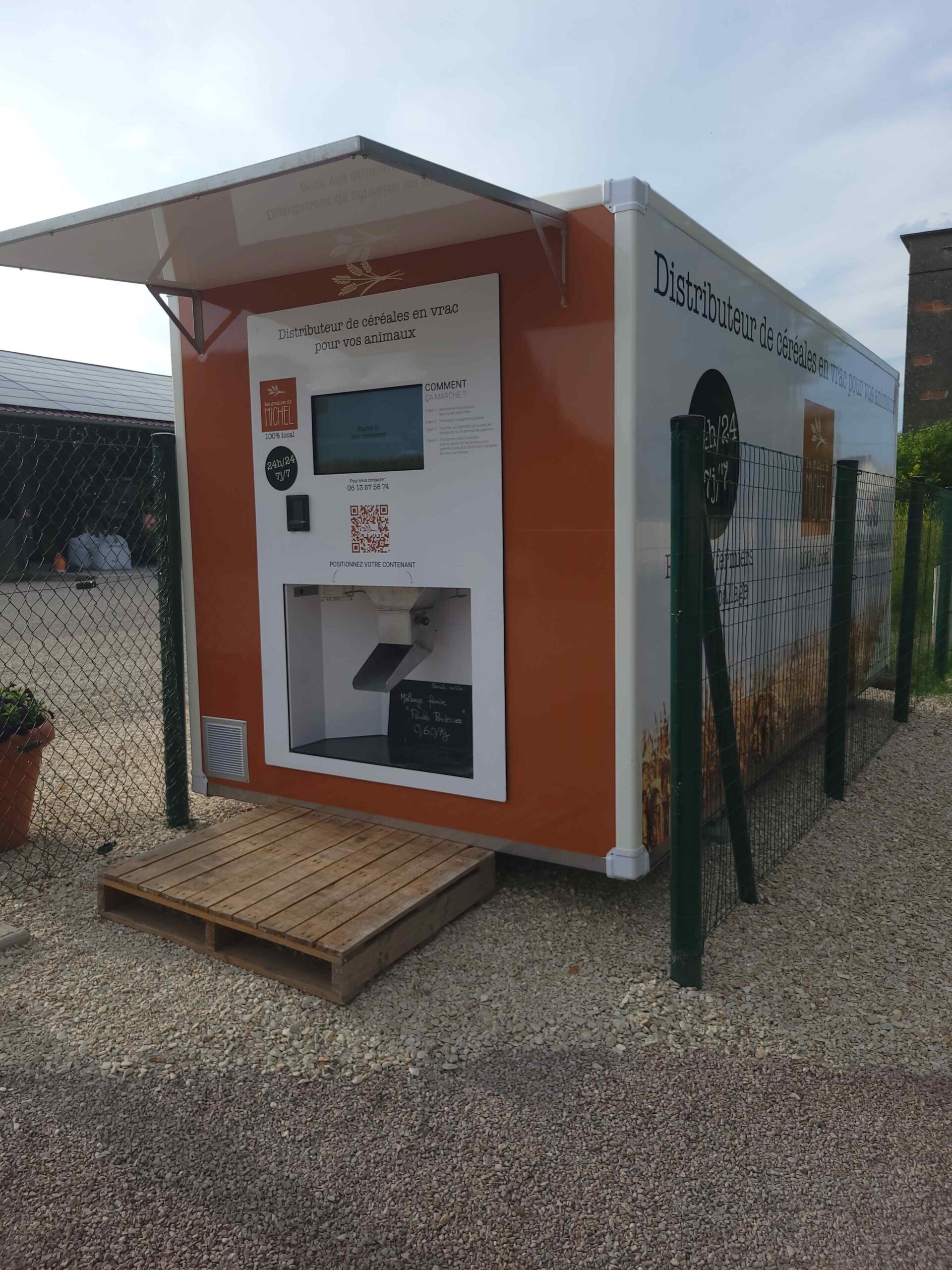 Distributeur de céréales en vrac pour animaux, extérieur, surface gravillonnée, toit protecteur.